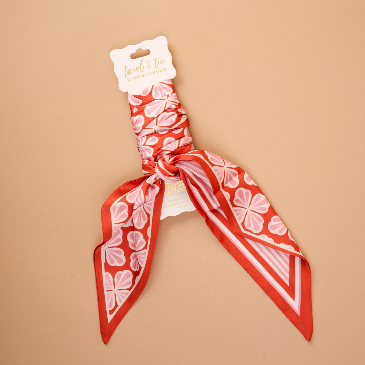 The Sienna Spice Twirl & Tie Scarf Accessory by The Darling Effect is a satin scarf in an eye-catching red and pink floral pattern, elegantly displayed against a beige backdrop. Featuring the brand’s signature label, a central knot, and pointed ends, this versatile accessory offers endless styling possibilities.