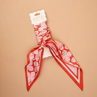 The Sienna Spice Twirl & Tie Scarf Accessory by The Darling Effect is a satin scarf in an eye-catching red and pink floral pattern, elegantly displayed against a beige backdrop. Featuring the brand’s signature label, a central knot, and pointed ends, this versatile accessory offers endless styling possibilities.