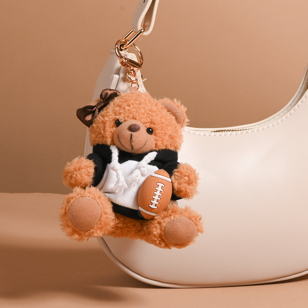 The Darling Effect’s Touchdown Teddy Teddy Bear Bag Charm—a small teddy bear with a brown bow, dressed in a black-and-white outfit and holding a plush football—adds playful style to any bag when attached to the handle of your white handbag, making it a perfect accessory or favorite charm against any beige backdrop.