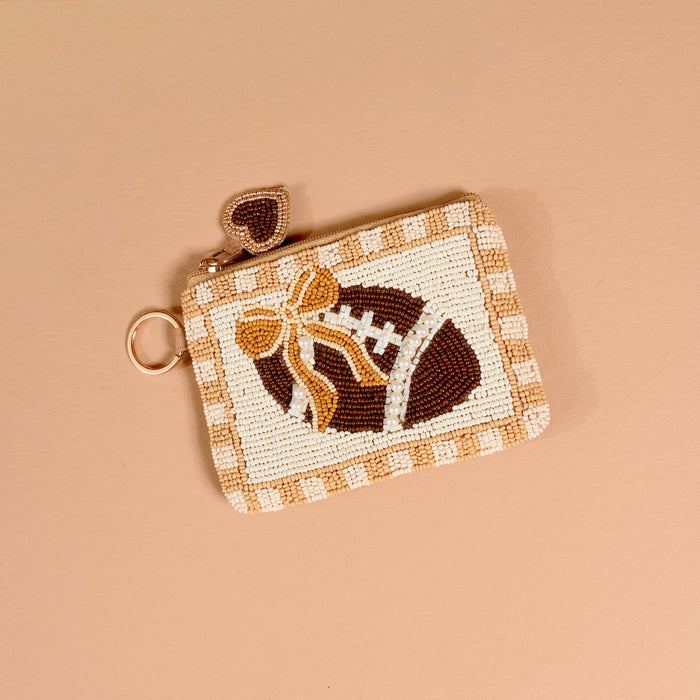 Introducing the Fab Football Handcrafted Beaded Pouch by The Darling Effect—a charming small pouch featuring a football design, embellished with a gold bow and finished with a heart-shaped zipper pull, displayed against a beige background.