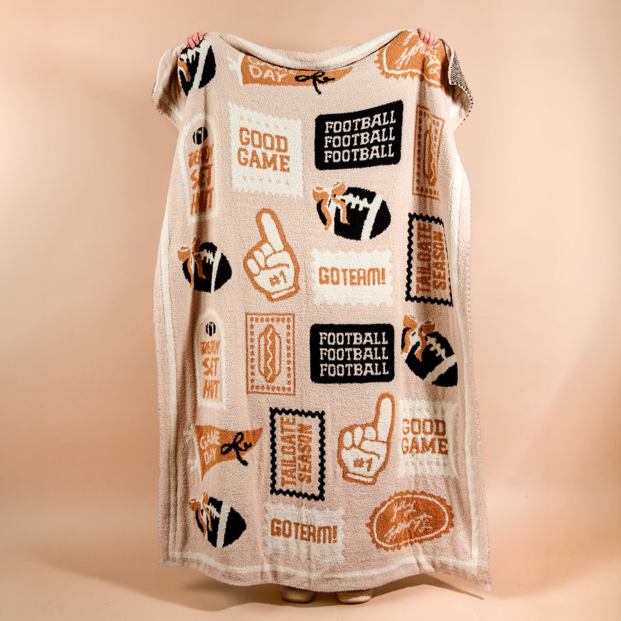A person holds up The Darling Effect Huddle Up Cozy Dreams Luxe Blanket, an ultra-soft beige blanket featuring football-themed designs like foam fingers, footballs, and playful phrases such as GOOD GAME, GOTEM!, and GAME DAY in brown, orange, and black. Only their feet are visible beneath this plush polyester microfiber blanket.