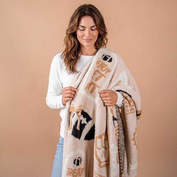 A woman with wavy brown hair in a white sweater smiles while holding The Darling Effect’s Huddle Up Cozy Dreams Luxe Blanket—a patterned beige and black, ultra-soft blanket—against a plain beige background.