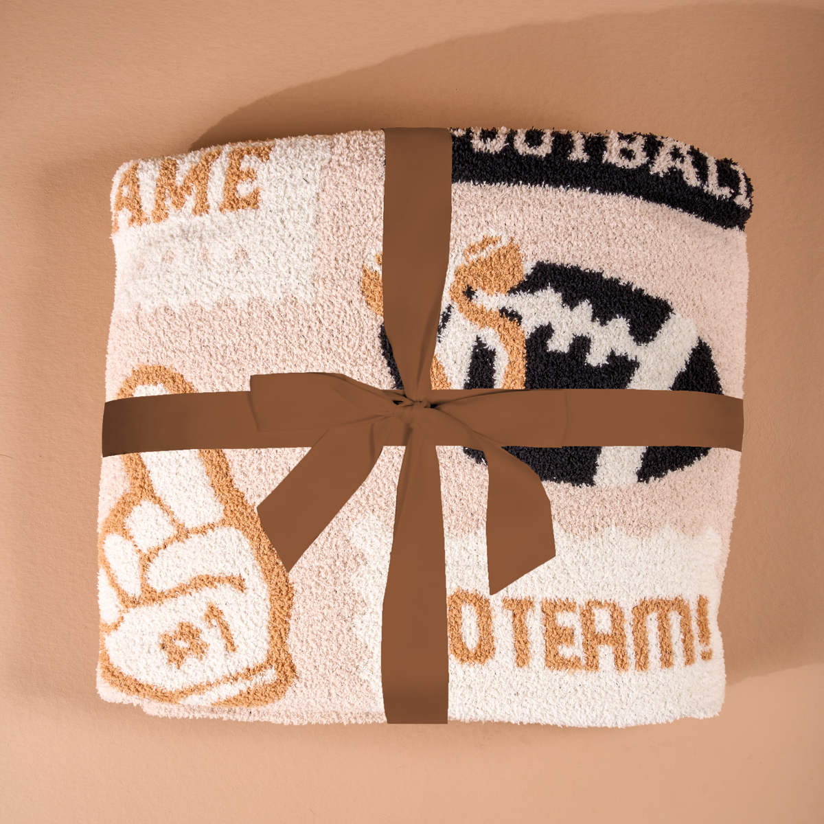 A folded Huddle Up Cozy Dreams Luxe Blanket by The Darling Effect, adorned with a brown ribbon, showcases sports-themed images like a foam finger and football along with partial words in beige and black on a light background. This ultra-soft blanket is displayed on a tan surface for an inviting presentation.