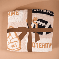 A folded Huddle Up Cozy Dreams Luxe Blanket by The Darling Effect, adorned with a brown ribbon, showcases sports-themed images like a foam finger and football along with partial words in beige and black on a light background. This ultra-soft blanket is displayed on a tan surface for an inviting presentation.