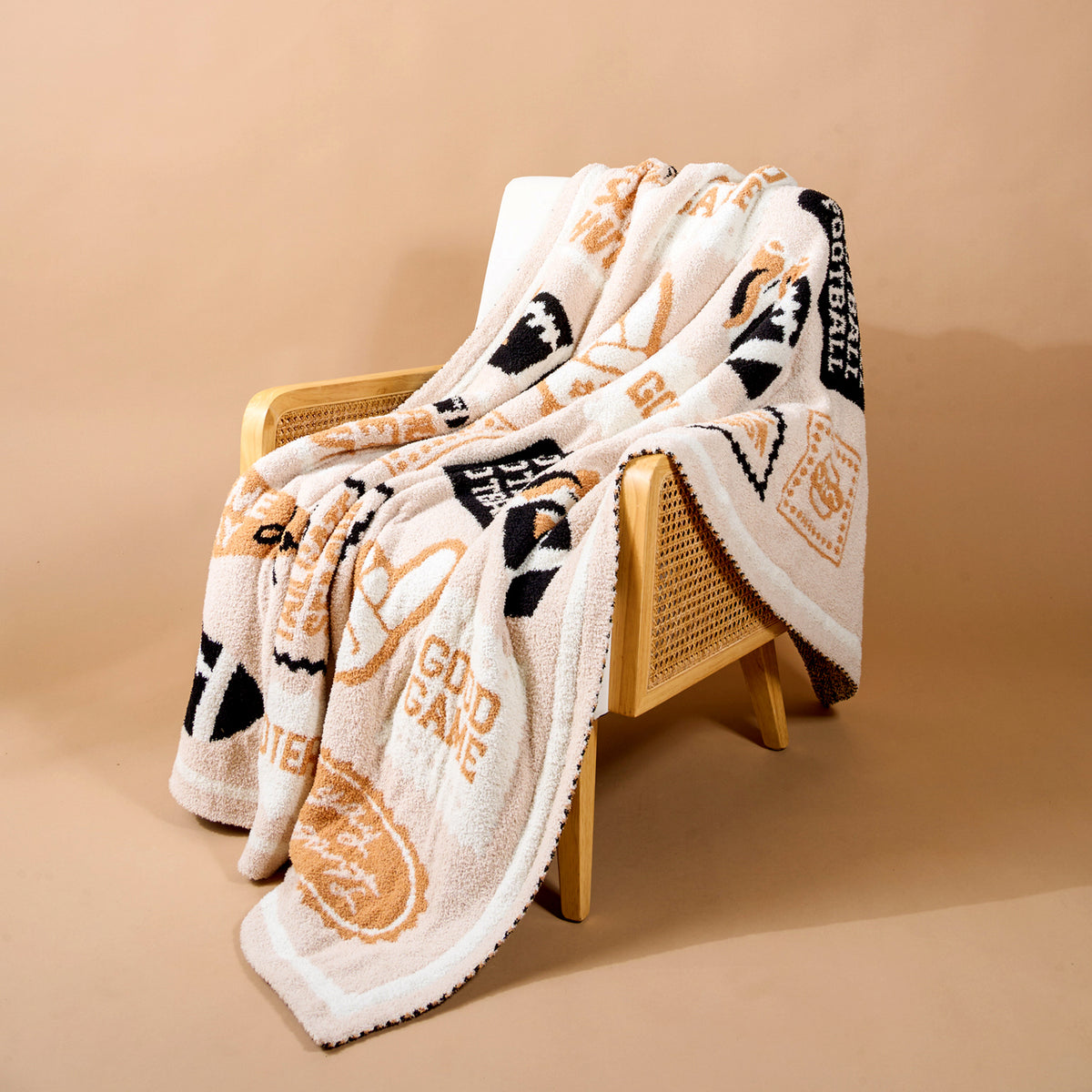 A cozy, patterned beige and white Huddle Up Cozy Dreams Luxe Blanket by The Darling Effect, featuring various text and graphic designs, drapes over a wooden chair with cane sides against a neutral tan background. This ultra-soft blanket is crafted from premium polyester microfiber for ultimate comfort.
