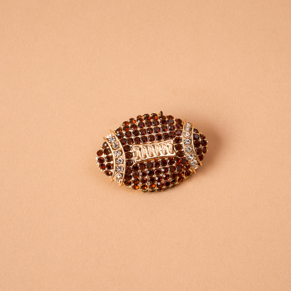 The Dazzle Football Timeless Brooch by The Darling Effect, a vintage-inspired accessory shaped like an American football and adorned with brown and clear rhinestones, rests on a plain beige background. This keepsake statement piece exudes classic charm and unique style.