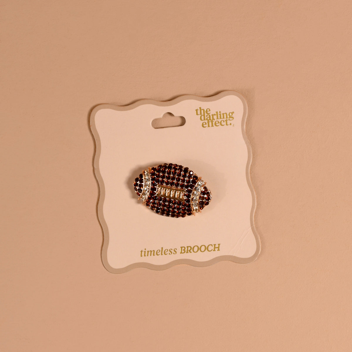 The Dazzle Football Timeless Brooch by The Darling Effect, featuring a vintage-inspired rhinestone football design, is attached to a card labeled “the darling effect. timeless BROOCH” and displayed against a beige background—making it a charming keepsake accessory.