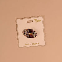 The Dazzle Football Timeless Brooch by The Darling Effect, featuring a vintage-inspired rhinestone football design, is attached to a card labeled “the darling effect. timeless BROOCH” and displayed against a beige background—making it a charming keepsake accessory.