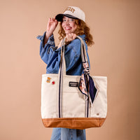 A smiling person in a cap and denim jacket holds The Darling Effect's Huddle Up The Darling Canvas Tote Bag, featuring brown leather accents, embroidered patches, a patterned scarf tied to the handle, and a spacious interior, standing against a plain beige background.