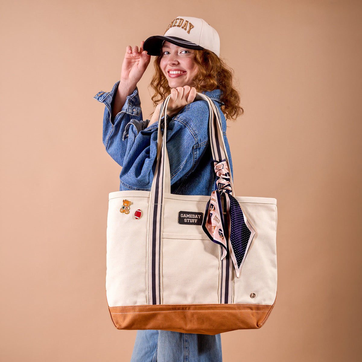 A smiling person in a denim jacket and The Darling Effect’s Gameday Classic Canvas Hat holds a large beige tote bag with brown trim, striped handles, and a scarf tied on. The bag is decorated with patches and pins. The background is a solid light brown color.