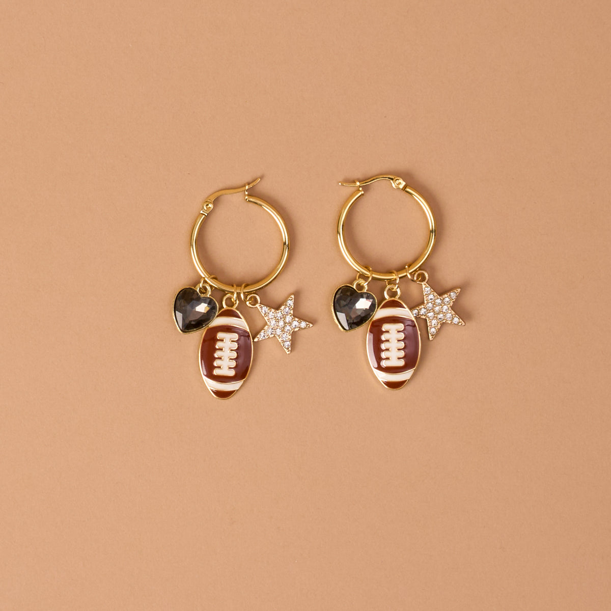 Gameday Glitz Signature Hoop Earrings by The Darling Effect feature gold hoops adorned with playful dangling charms—a brown football, a black heart, and a silver star embellished with rhinestones—against a simple tan background. These lightweight earrings offer the perfect mix of fun and elegance, making them ideal for any occasion.