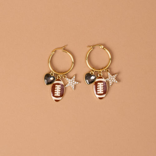 Gameday Glitz Signature Hoop Earrings by The Darling Effect feature gold hoops adorned with playful dangling charms—a brown football, a black heart, and a silver star embellished with rhinestones—against a simple tan background. These lightweight earrings offer the perfect mix of fun and elegance, making them ideal for any occasion.