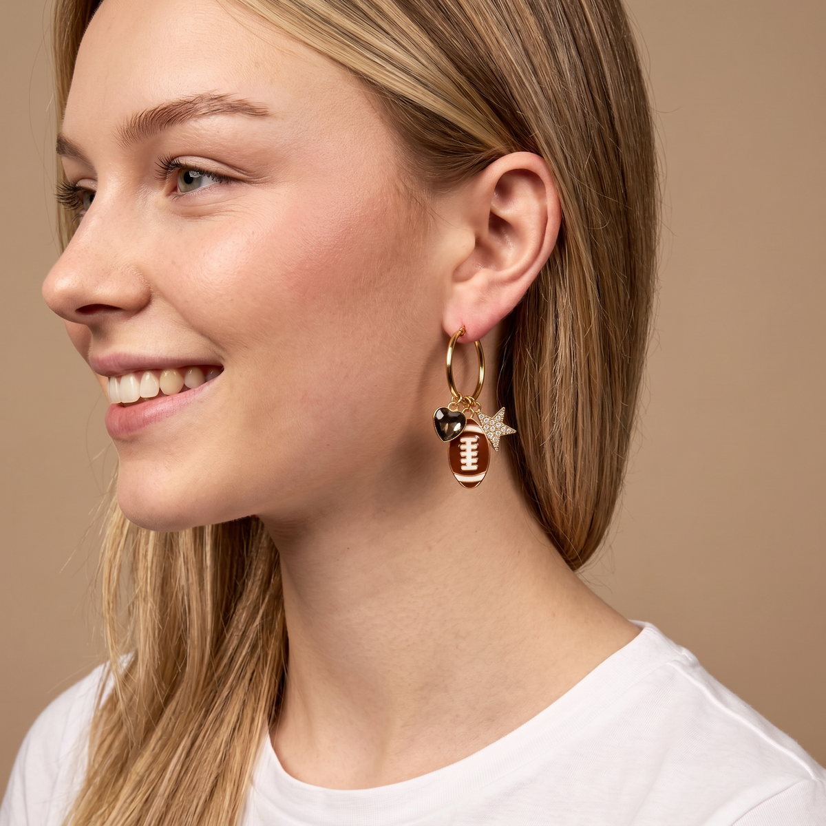 A smiling woman with straight blonde hair wears a white shirt and The Darling Effect's Gameday Glitz Signature Hoop Earrings—lightweight gold hoops featuring football and goalpost charms—against a beige background.