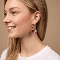 A smiling woman with straight blonde hair wears a white shirt and The Darling Effect's Gameday Glitz Signature Hoop Earrings—lightweight gold hoops featuring football and goalpost charms—against a beige background.