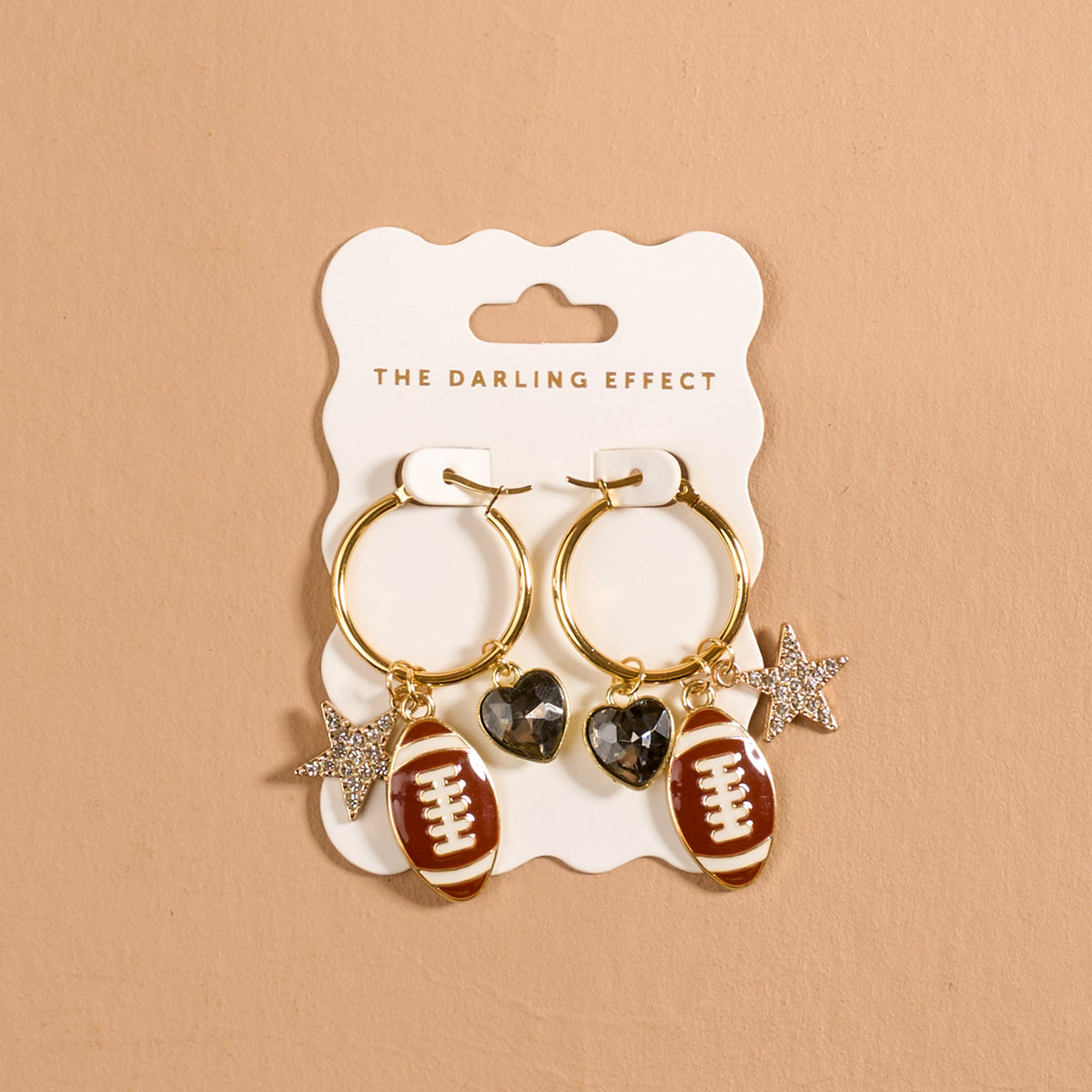 The Gameday Glitz Signature Hoop Earrings by The Darling Effect feature two lightweight gold hoops adorned with football, heart, and star charms, displayed on a branded white card against a beige background—chic charm earrings that effortlessly elevate any look.