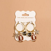 The Gameday Glitz Signature Hoop Earrings by The Darling Effect feature two lightweight gold hoops adorned with football, heart, and star charms, displayed on a branded white card against a beige background—chic charm earrings that effortlessly elevate any look.