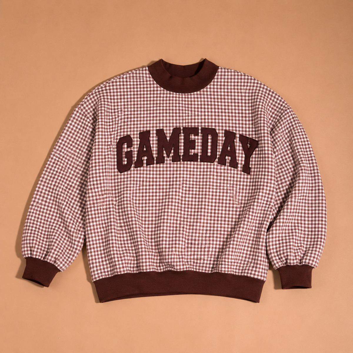 The Gameday Mockneck Sweatshirt by The Darling Effect features a brown and cream checkered design with “GAMEDAY” in bold brown letters across the front, shown against a solid tan background.
