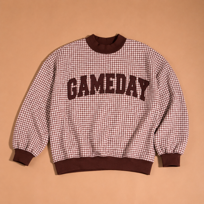 The Gameday Mockneck Sweatshirt by The Darling Effect features a brown and cream checkered design with “GAMEDAY” in bold brown letters across the front, shown against a solid tan background.