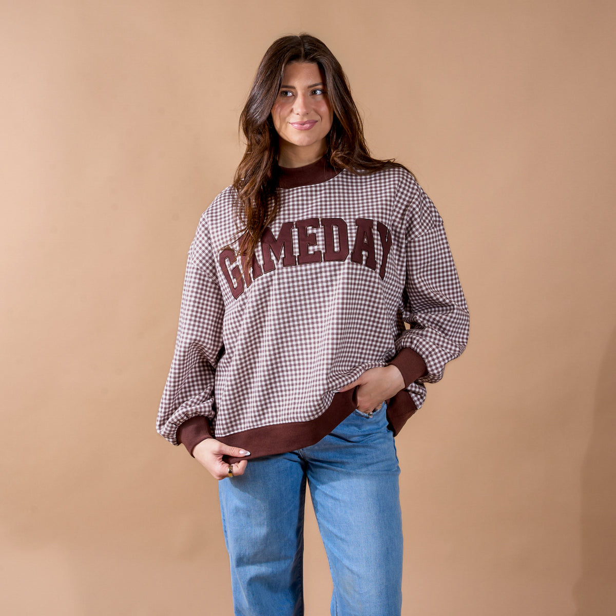 A woman wearing The Darling Effect's Gameday Mockneck Sweatshirt—a brown, checkered top with "GAMEDAY" on the front—paired with blue jeans stands against a plain tan background, smiling slightly.