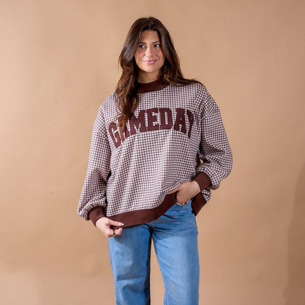 A woman wearing The Darling Effect's Gameday Mockneck Sweatshirt—a brown, checkered top with "GAMEDAY" on the front—paired with blue jeans stands against a plain tan background, smiling slightly.