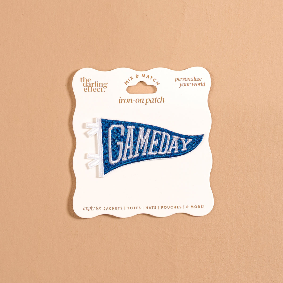 A blue pennant-shaped iron-on patch reading “GAMEDAY” in white letters from The Darling Effect, the Gameday Blue Mix & Match Iron-On Patch is presented on cream packaging against a beige background. Ideal for personalizing items like jackets, totes, hats, and more, as suggested on the packaging.