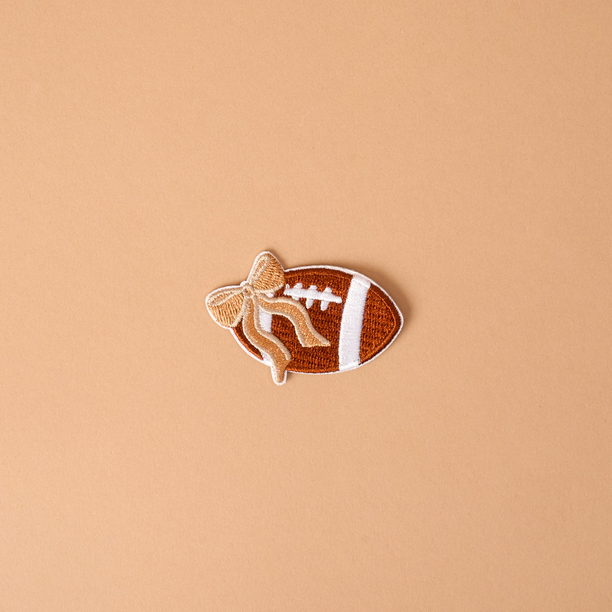 Introducing the Gameday Fab Football Mix & Match Iron-On Patch by The Darling Effect—a football-shaped patch featuring white laces and a tan bow, set against a plain light brown background. Perfect for anyone wanting to personalize items with this unique, easy-to-use iron-on accessory.