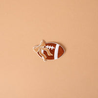 Introducing the Gameday Fab Football Mix & Match Iron-On Patch by The Darling Effect—a football-shaped patch featuring white laces and a tan bow, set against a plain light brown background. Perfect for anyone wanting to personalize items with this unique, easy-to-use iron-on accessory.
