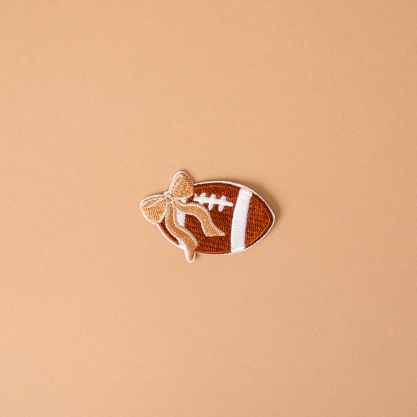 Introducing the Gameday Fab Football Mix & Match Iron-On Patch by The Darling Effect—a football-shaped patch featuring white laces and a tan bow, set against a plain light brown background. Perfect for anyone wanting to personalize items with this unique, easy-to-use iron-on accessory.