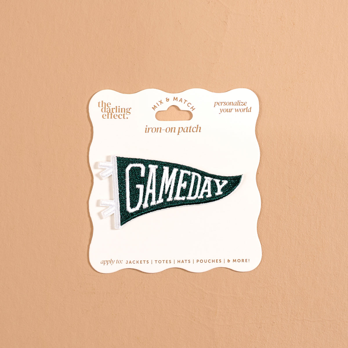 The Darling Effect’s Gameday Green Mix & Match Iron-On Patch features a green pennant shape with bold white “GAMEDAY” lettering, attached to a beige card labeled “iron-on patch” and “personalize your world.” Perfect for anyone looking to customize their gear, this patch is showcased against a neutral tan background.
