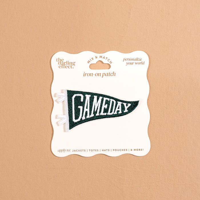 The Darling Effect’s Gameday Green Mix & Match Iron-On Patch features a green pennant shape with bold white “GAMEDAY” lettering, attached to a beige card labeled “iron-on patch” and “personalize your world.” Perfect for anyone looking to customize their gear, this patch is showcased against a neutral tan background.