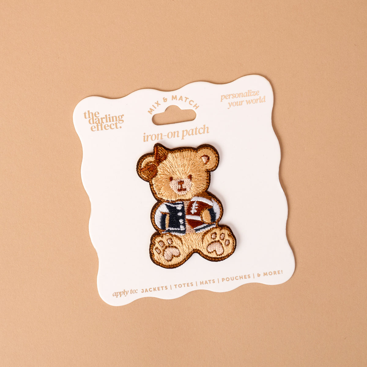 The Gameday Touchdown Teddy Mix & Match Iron-On Patch by The Darling Effect features a teddy bear in a letterman jacket, displayed on a white card with wavy edges against a beige background. Part of our colorful patch collection, this vibrant patch is perfect for adding a playful touch to jackets, totes, hats, and pouches.