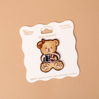 The Gameday Touchdown Teddy Mix & Match Iron-On Patch by The Darling Effect features a teddy bear in a letterman jacket, displayed on a white card with wavy edges against a beige background. Part of our colorful patch collection, this vibrant patch is perfect for adding a playful touch to jackets, totes, hats, and pouches.