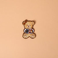 The Gameday Touchdown Teddy Mix & Match Iron-On Patch by The Darling Effect features an embroidered brown teddy bear dressed in a blue jacket and holding a small brown item, set against a plain beige background—ideal for anyone who loves customizable patches or wants to add unique iron-on designs to their collection.