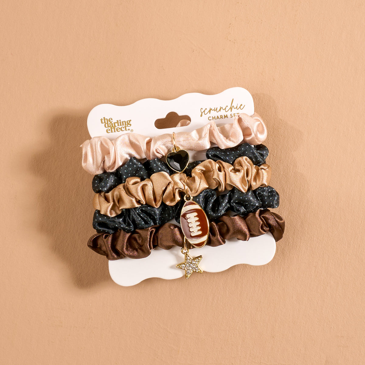 Introducing the Tailgate Season Scrunchie Charm Set by The Darling Effect, featuring five chic hair accessories in beige, black, and brown tones. Each scrunchie is elegantly arranged on a display card and adorned with two charming accents—a gold star and a football—for a playful touch. The set is showcased against a simple light brown background.