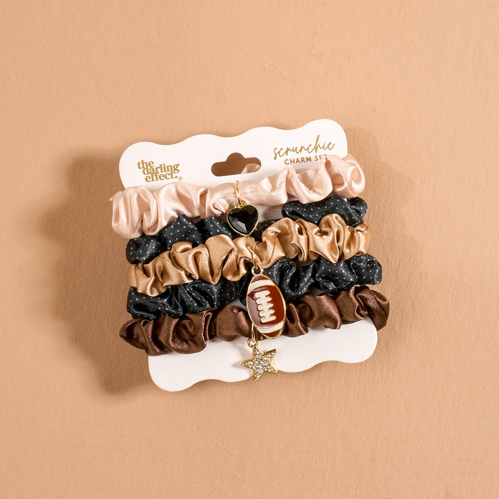 Introducing the Tailgate Season Scrunchie Charm Set by The Darling Effect, featuring five chic hair accessories in beige, black, and brown tones. Each scrunchie is elegantly arranged on a display card and adorned with two charming accents—a gold star and a football—for a playful touch. The set is showcased against a simple light brown background.