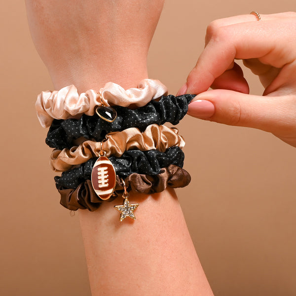 A hand wears The Darling Effect's Tailgate Season Scrunchie Charm Set—stylish beige, black, and brown scrunchies, each accented with playful football, star, jewel, and heart charms—against a plain brown background. These charming hair accessories add a touch of playful elegance to any look.