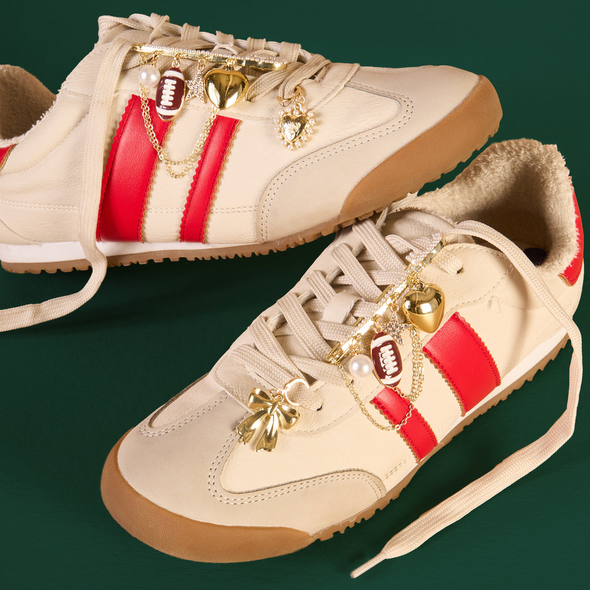 Two beige sneakers with red stripes and gum soles showcase The Darling Effect’s Tailgate Time Dazzle Shoe Charm Pack—featuring football, heart, pearl, and other fun charms—adding style against a dark green background.