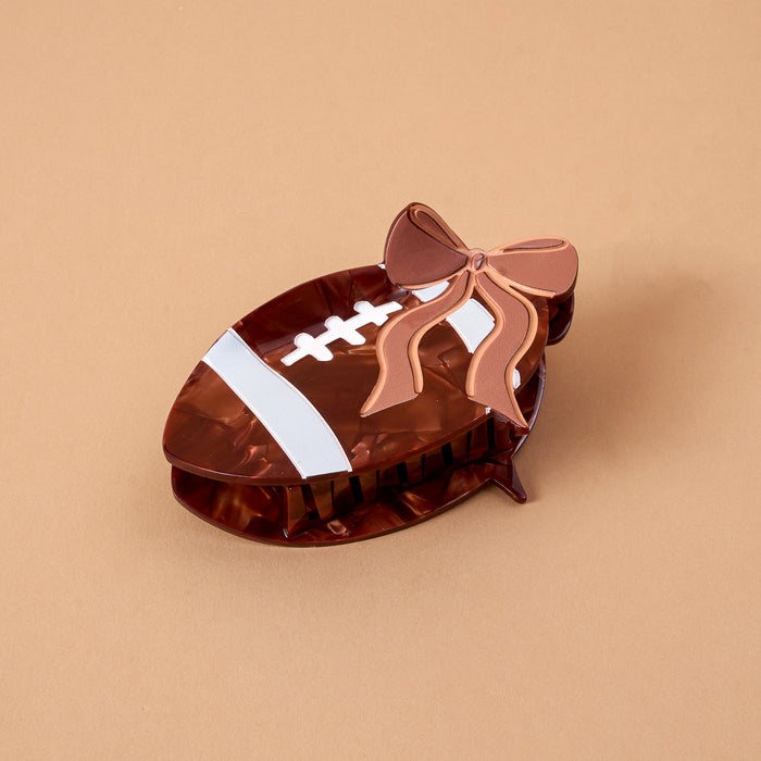 The Darling Effect’s Fab Football Shape Up Claw Clip, designed in a playful football shape with delicate white lace accents and a pink and brown decorative bow, rests elegantly on a beige surface. This charming hair accessory is perfect for adding a fun and stylish touch to any hairstyle.