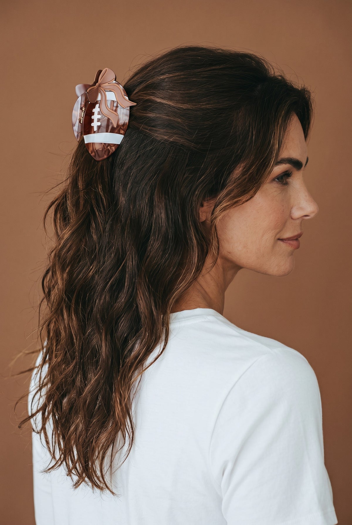 A woman with wavy brown hair, dressed in a white shirt, styles her hair with The Darling Effect's Fab Football Shape Up Claw Clip as she stands against a brown background.
