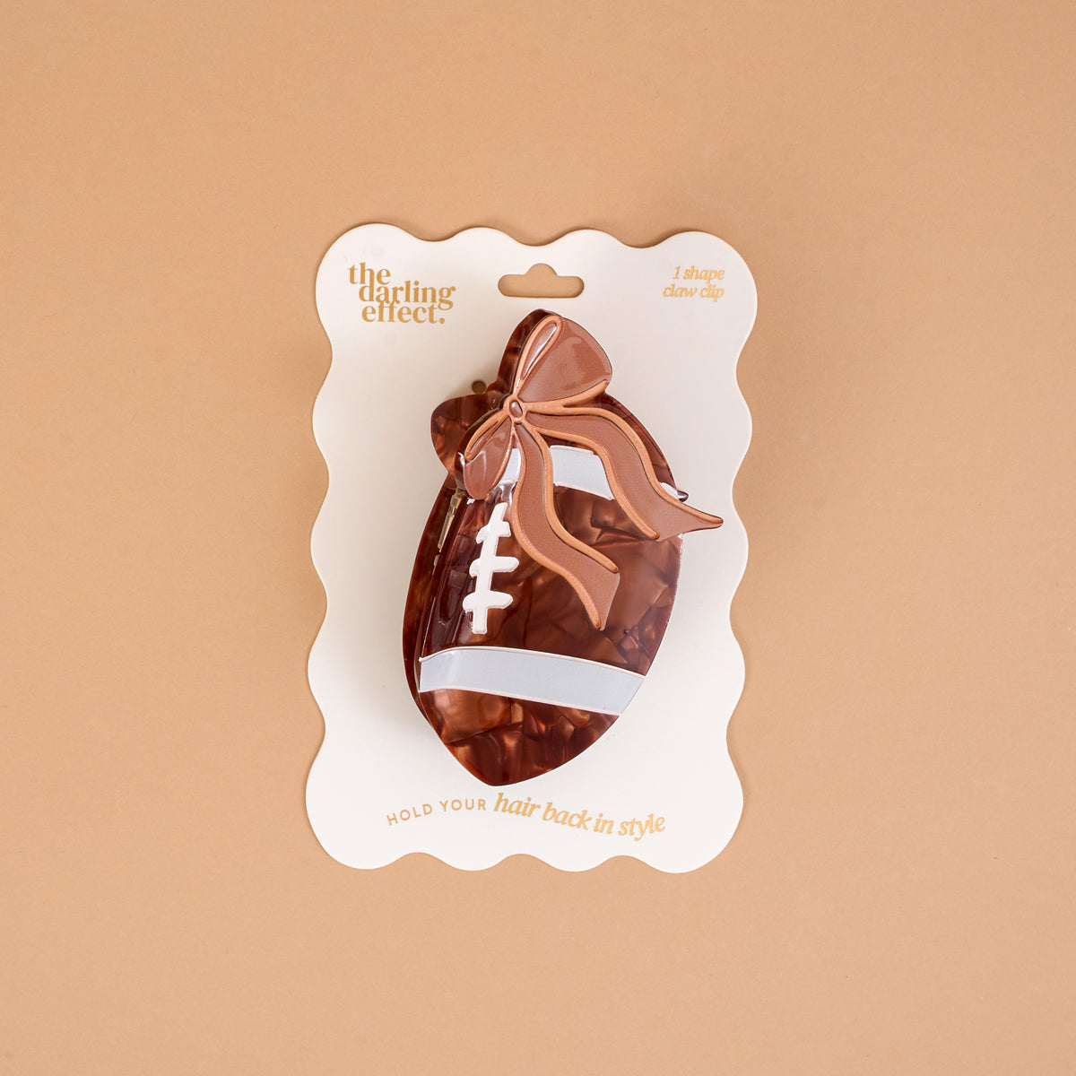 Introducing the Fab Football Shape Up Claw Clip by The Darling Effect—a charming brown hair accessory featuring a football shape with an adorable bow, beautifully presented on branded packaging against a beige background.