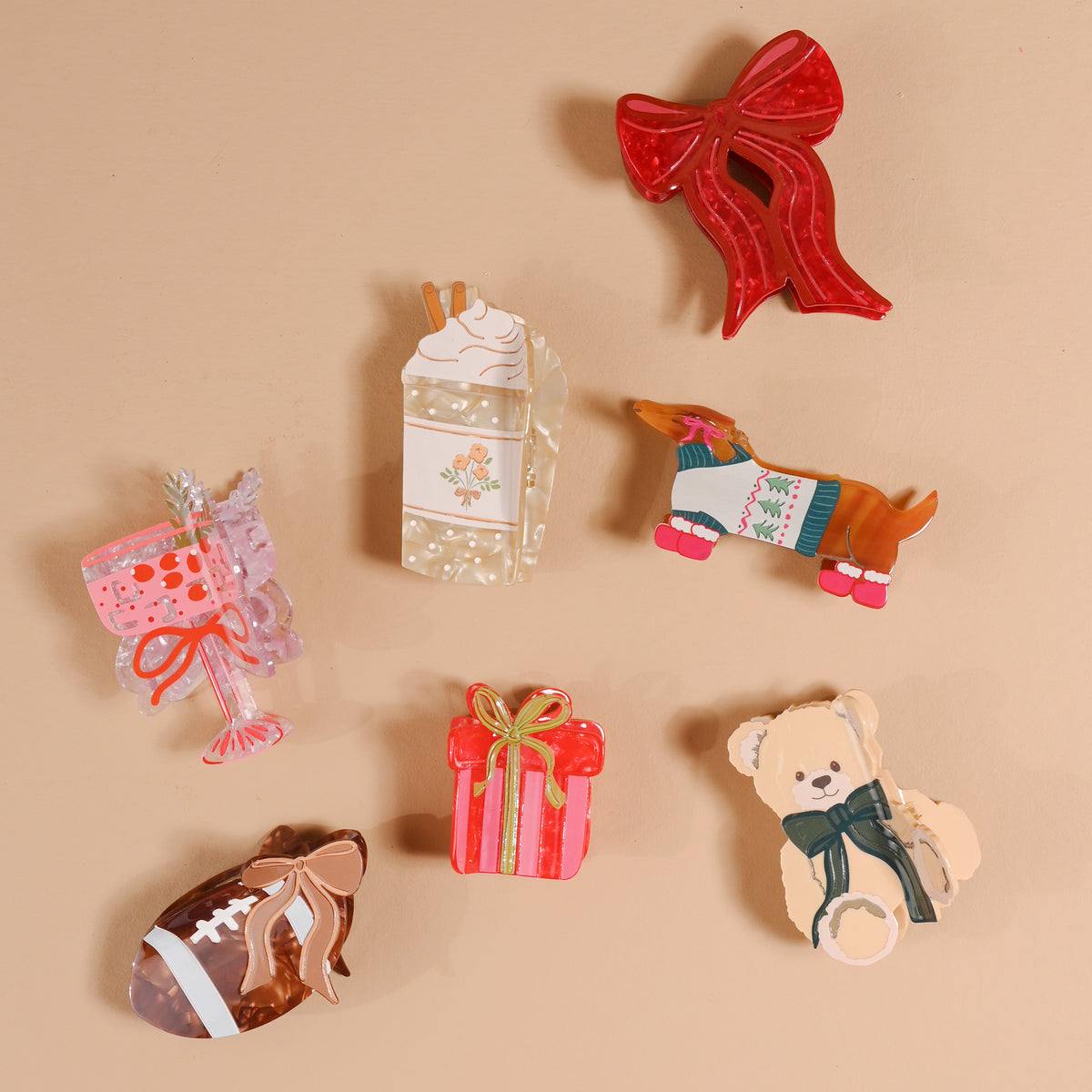 Seven holiday-themed pins—including a red bow, a milk carton, a sausage dog in a sweater, a pink dessert, a wrapped gift, a teddy bear with a bow, and a brown football—are arranged on a beige background. Each pin is as delightful as The Darling Effect’s Fab Football Shape Up Claw Clip and makes the perfect festive accent for your collection or to complement your favorite hair accessories.