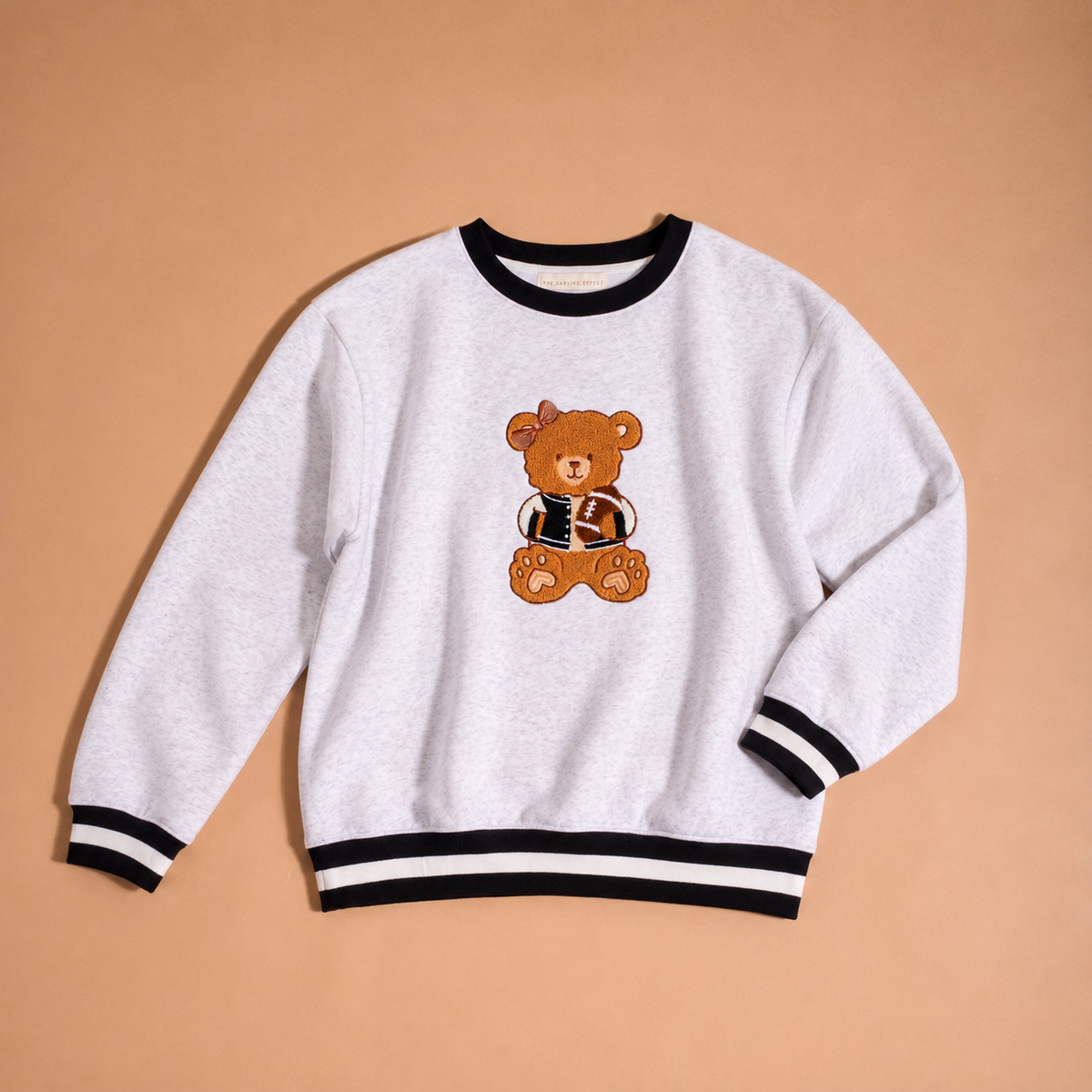 The Darling Effect Touchdown Teddy Signature Crew is a light gray sweatshirt with black and white striped cuffs and hem, featuring an embroidered brown teddy bear in a jacket on the front, displayed on a tan background.