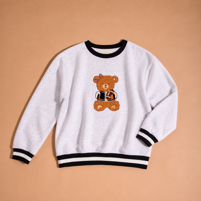 The Darling Effect Touchdown Teddy Signature Crew is a light gray sweatshirt with black and white striped cuffs and hem, featuring an embroidered brown teddy bear in a jacket on the front, displayed on a tan background.
