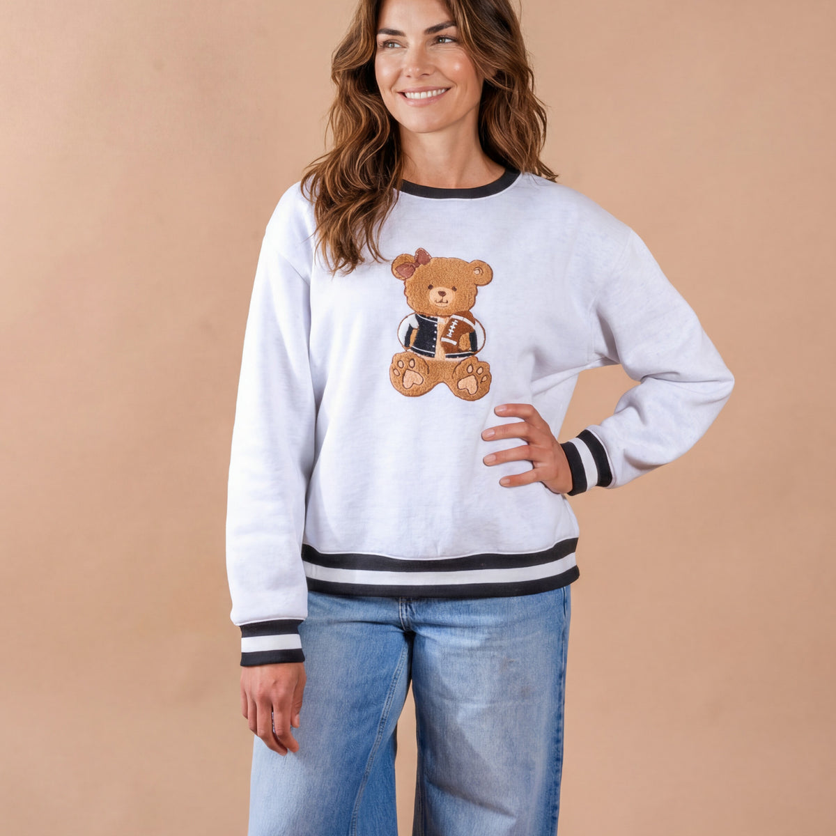 A woman wearing The Darling Effect's Touchdown Teddy Signature Crew and blue jeans stands smiling against a plain beige background.