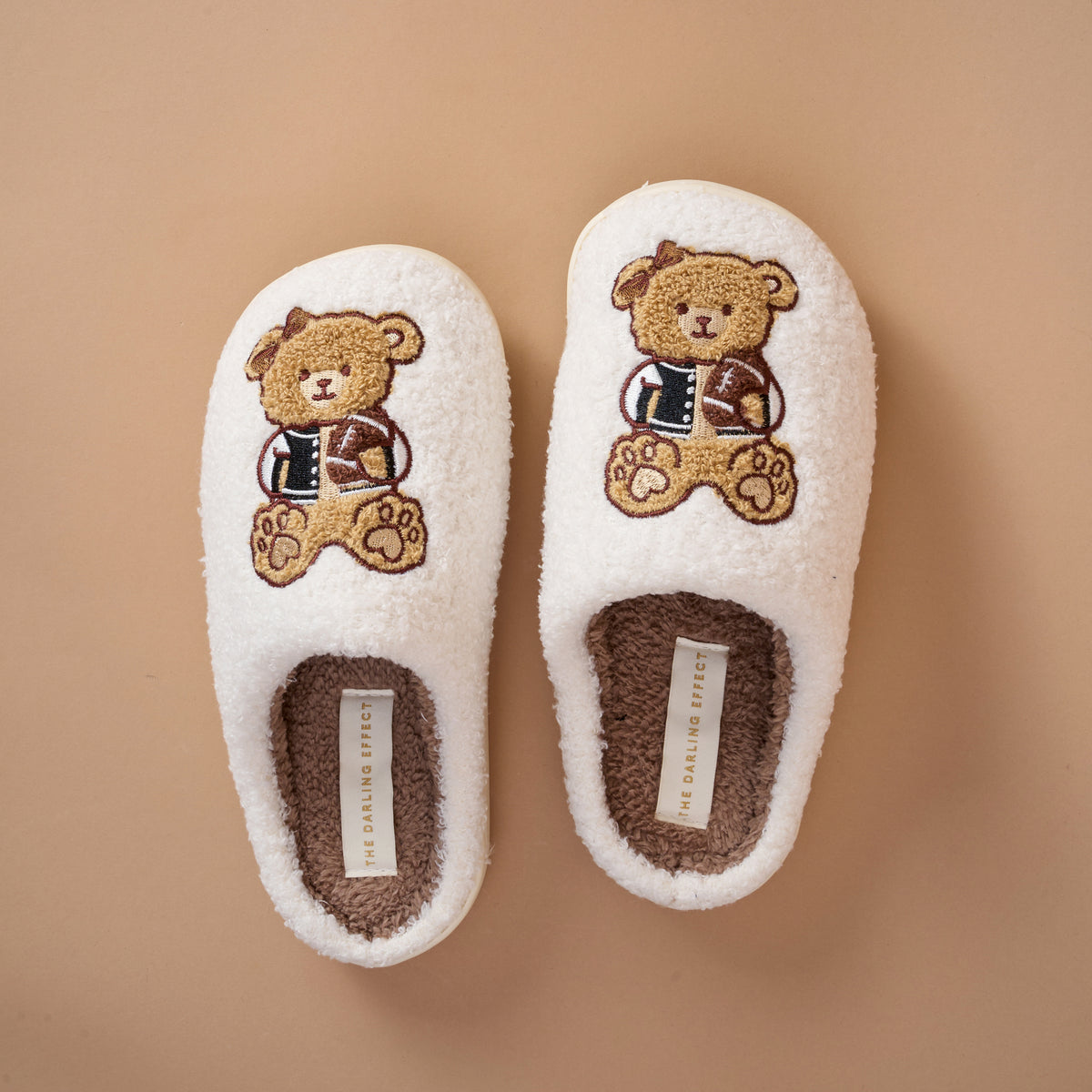 A pair of Touchdown Teddy Fuzzy Slippers by The Darling Effect, in white with brown soles and embroidered teddy bears wearing sweaters, are placed side by side on a beige background.
