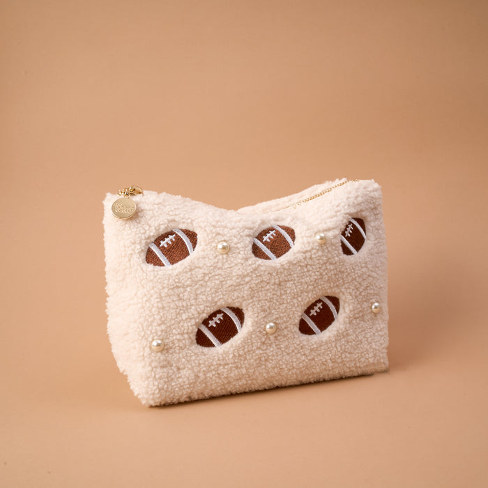The Football & Pearls Teddy Pouch by The Darling Effect features a fuzzy, cream-colored sherpa zip pouch adorned with delicate pearl accents and playful footballs peeking through cutout details, displayed on a simple beige background.