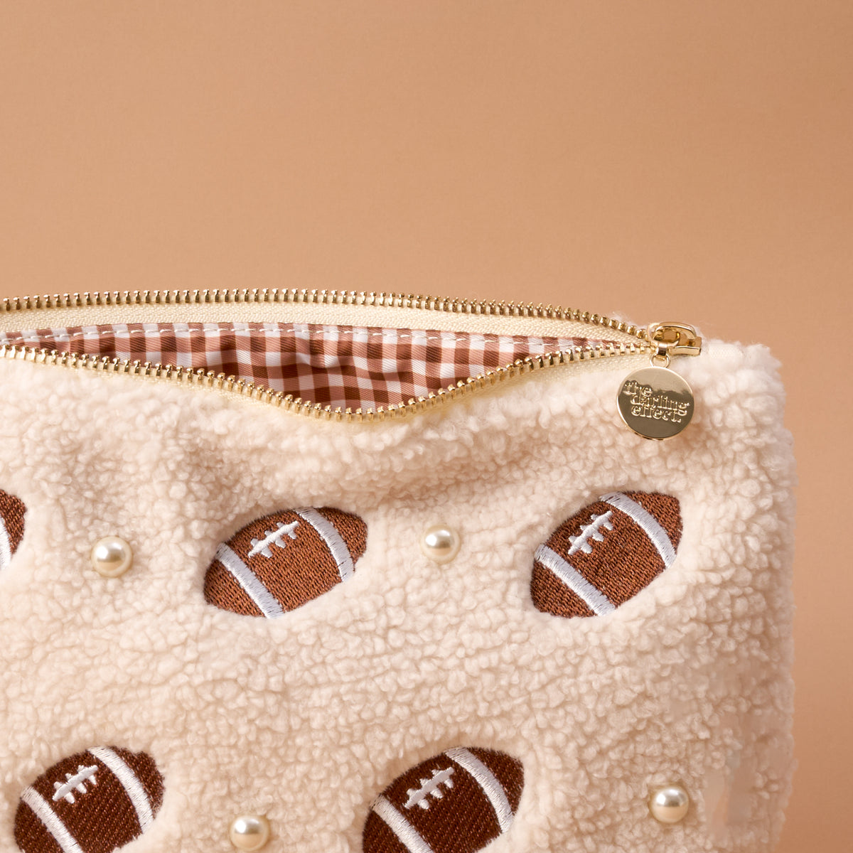 Introducing the Football & Pearls Teddy Pouch by The Darling Effect—a cozy, cream-colored zip pouch made from plush sherpa fabric. This charming accessory features embroidered footballs, delicate pearls, a gleaming gold zipper with a small round charm inscribed with text, and a classic gingham lining. Set against a solid beige background, it’s perfect on its own or paired with other teddy pouches as part of your accessory collection.