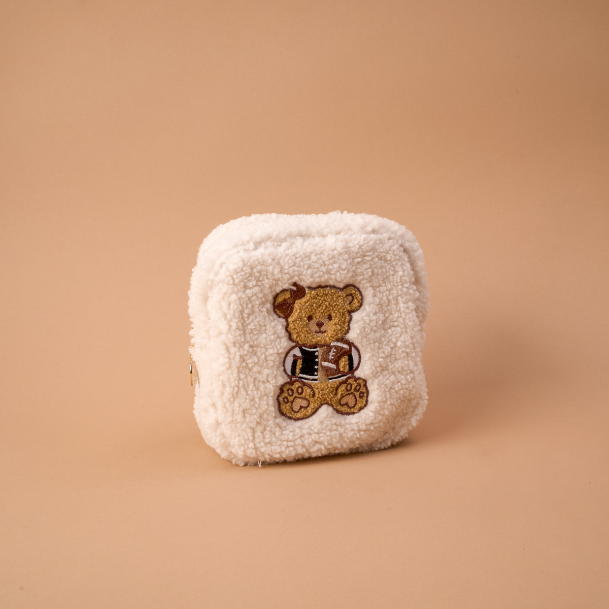 The Square Touchdown Teddy Teddy Pouch by The Darling Effect is a small, square pouch made from soft, cream-colored sherpa fabric, featuring an embroidered teddy bear in a jacket on the front and displayed against a plain beige background.