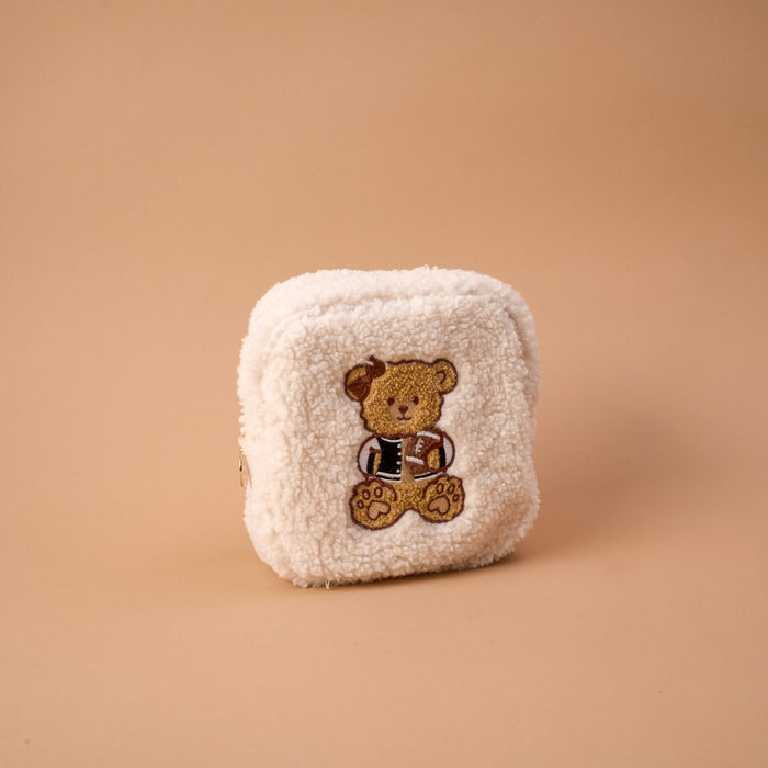 The Square Touchdown Teddy Teddy Pouch by The Darling Effect is a small, square pouch made from soft, cream-colored sherpa fabric, featuring an embroidered teddy bear in a jacket on the front and displayed against a plain beige background.