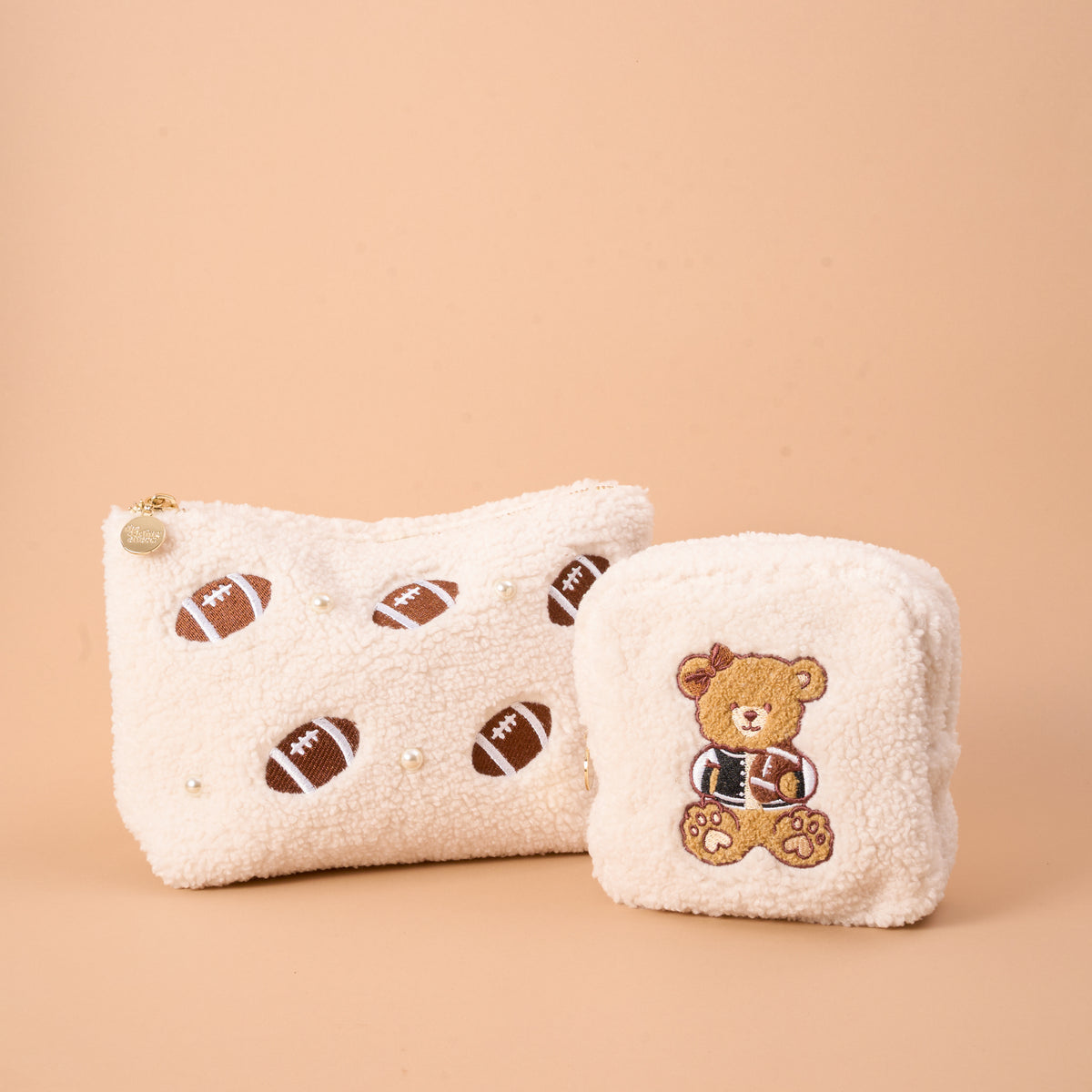 Add a playful touch to your collection with The Darling Effect's Square Touchdown Teddy Teddy Pouches—two soft sherpa pouches in creamy beige, including one embroidered with footballs and pearl accents, and another showcasing an adorable teddy bear holding a football.